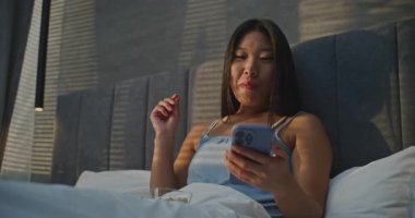 Güzel Asyalı kadın yatakta fındık yiyor ve internette arkadaşlarıyla sohbet ediyor. Kız İnternette Ceviz ve İletişim 'i seviyor. Sabah Rutini Konsepti, Yatakta Kahvaltı, Dijital Hayat. El bilgisayarı.