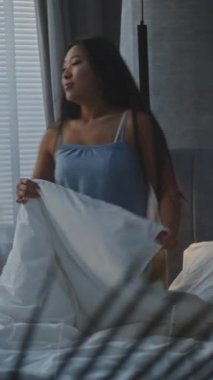 Şık Yatak Odası 'ndaki Uzun Saçlı Asyalı Kız Yatakta Rahat Uzanıyor, Üstünden Battaniyeyi Çekiyor, Smartphone' da Çevrimiçi Hayata Batıyor. Sabah Rutini kavramı, Dijital Yaşam Tarzı. Dikey Çekim.