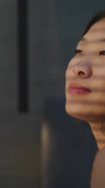 Genç Kadın Güneşli Yatak Odasında Sabah Uyanır. Kız uykulu bir gülümsemeyle yatakta oturur, pencereden dışarı bakar, güneş ışığı yüzüne vurur. Günaydın, Pozitif Ruh hali. Rahatlamayı kabul et, uyu. Dikey Çekim.