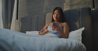 Genç Kadın Sabahları Yatakta Kahvaltı Yiyor, Sosyal Medya Akıllı Telefonla Besleniyor. Kızlar sandviçi sever, sohbetler internetten. Sabah Rutini Konsepti, Yatakta Kahvaltı, Dijital Yaşam Tarzı.