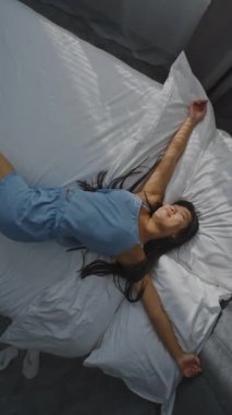 Asyalı Kız Pamuk Prenses 'in üzerine düşüyor. Kolları açık, Gülümseyen Mutlulukla, Gerinen, Yatakta Parlayan. Rahatlama, rahatlama, güzel bir sabah, sağlıklı bir yaşam tarzı. Dönen Üst Görünüm. Dikey Çekim.