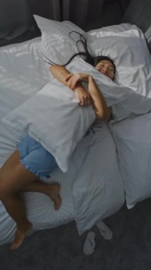 Sunlit Yatak Odası 'nda, Genç Asyalı Esmer Kız Beyaz Yatakta Bass yapıyor. Güzel kadın gülümsüyor, yastığa sarılıyor. Rahatlama, Akıl Sağlığı, Olumlu Duygular ve Keyif. Dönen Üst Görünüm. Dikey Çekim.