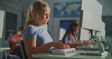 İlkokul öğrencileri Parlak Sınıfta Öğrenir. Mavi Gömlekli Sarışın Kız, Sınıf Arkadaşları Masada Otururken Masaüstü Bilgisayarı 'nda çalışıyor. E-öğrenim kavramı, çocukluk gelişimi, çalışma. El bilgisayarı.