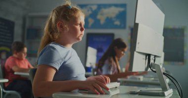 İlkokul öğrencileri Parlak Sınıfta Öğrenir. Mavi Gömlekli Sarışın Kız, Sınıf Arkadaşları Masada Otururken Masaüstü Bilgisayarı 'nda çalışıyor. E-öğrenim kavramı, çocukluk gelişimi, çalışma. El bilgisayarı.
