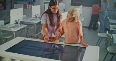 Parlak Sınıfta İki Liseli Kız Büyük Etkileşimli Dokunmatik Masa Kullanır. Çocuklar Eğitimsel İçeriği Merakla Keşfeder. Dijital Öğrenme ve Modern Okul Teknolojisi kavramı. El bilgisayarı.