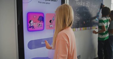 Sınıftaki Genç Sarışın Kız Öğrenci Dijital Ekranda Temperament Testi Yapıyor. Diğer öğrenciler tahtaya yazı yazıyor. Duygusal Zeka, EdTech ve Öğrenci Refahı kavramı. Arka Görünüm.