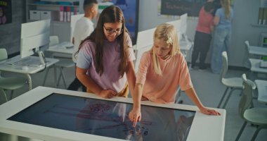 Parlak Sınıfta İki Liseli Kız Büyük Etkileşimli Dokunmatik Masa Kullanır. Çocuklar Eğitimsel İçeriği Merakla Keşfeder. Dijital Öğrenme ve Modern Okul Teknolojisi kavramı..