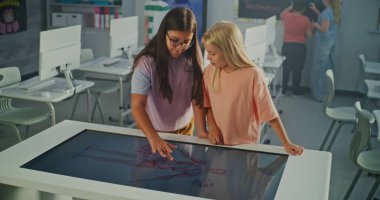 Parlak Sınıfta İki Liseli Kız Büyük Etkileşimli Dokunmatik Masa Kullanır. Çocuklar Eğitimsel İçeriği Merakla Keşfeder. Dijital Öğrenme ve Modern Okul Teknolojisi kavramı. El bilgisayarı.