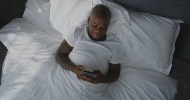 Yetişkin Adam, Smartphone Haber Kaynağını Sabah Yatağında Geçiriyor. Gülümseyen Afrikalı Amerikalı Adam Sosyal Medyadan İçerik Bakıyor. Concept Connected Lifestyle, Morning Digital Routine. Üst Görünüm Döndürülüyor.