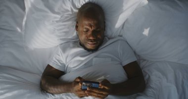 Adam Yoğun Güneş Işığından Yavaşça Uyanır ve Akıllı Telefona Ulaşır. Yakışıklı Afrikalı Amerikalı, Güneş 'ten İnekleyen, Çevrimiçi Sabah Programıyla Başlıyor. Bağlı Yaşam Tarzı. Sabit Çekim.