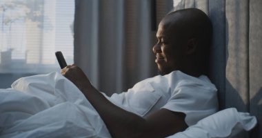 Huzurlu Sabah Anı Akıllı Telefonlu Yatakta Odaklanan Adam, Gülümseyen Ekran. Yetişkin Adam Yatak Odasında, Güneş Işığı Perdelerin Arasında. Kombinasyon Teknolojisi, Rahatlama, Rahatlık ve Pozitif Ruh hali.