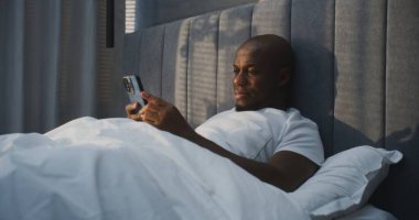 Beyaz Pijamalı Gülümseyen Adam Yatakta Rahatlıyor, Akıllı Telefonu Ellerinde Tutuyor. Sıcak Sabah Işığında Neşeli Adam Yazıyor ve Okuyor. Concept Digital Lifestyle, Cozy Rest ve Online Connection. El bilgisayarı.