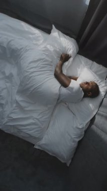 Afro-Amerikan Adam Sabah Uyanır ve Parlak Güneş Işığında İnekler. Yetişkin adam uyandıktan sonra beyaz yatakta gülümser ve gerilir. Rahatlama, dinlenme, sağlıklı uyku. Dönen Üst Görünüm. Dikey Çekim.