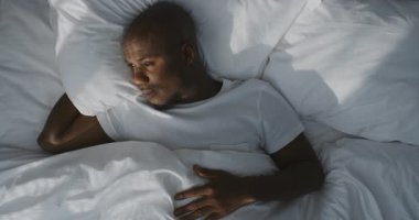 Beyaz pijamalı Barışçıl Adam Kar Beyaz Yatağında Yavaşça Uyanıyor, Sıcak Güneş Işığıyla Banyo Yapıyor. Rahatlama kavramı, sağlıklı yaşam tarzı, rahat bir sabah rutini, konfor ve dinlenme. Sabit Çekim.