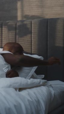 Sabah Yakışıklı Afro-Amerikan Adam, Alarm Saatinin Yatak Sesi 'nde Uyanıyor. Adam yataktan çıkıyor, parlak güneş ışığına karşı gözlerini kısarak bakıyor. Kavram Sabah Uyanışı, her sabah. Dikey Çekim.