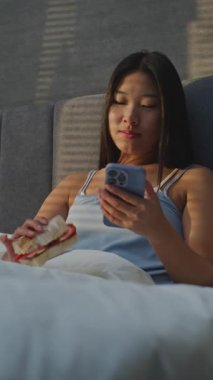 Genç Kadın Sabahları Yatakta Kahvaltı Yiyor, Sosyal Medya Akıllı Telefonla Besleniyor. Kızlar sandviçi sever, sohbetler internetten. Sabah rutini, Yatakta Kahvaltı, Dijital Yaşam Tarzı. Dikey Çekim.