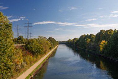 Nehir ağaçların arasından akar. Ufuktaki elektrik kuleleri. Güneşli bir gün ve gökyüzünde bulutlar.