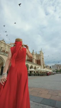 Kırmızı elbiseli bir kadın, üzerinde güvercinler uçarken ana meydanda yürüyor ve kıvrılıyor..