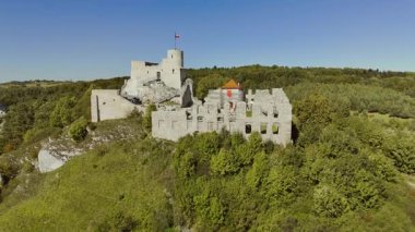 İnsansız hava aracı, Rabsztyn Kalesi 'ni yukarıdan yakalıyor, ortaçağ taş duvarlarını ve kulelerini vurguluyor, etrafını çevreleyen orman ve manzaralarla çevrili bir tepenin üzerine kuruluyor..