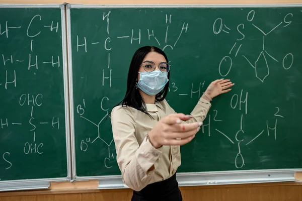 young female teacher in protective mask standing near blackboard. chemical formulas written on classboard