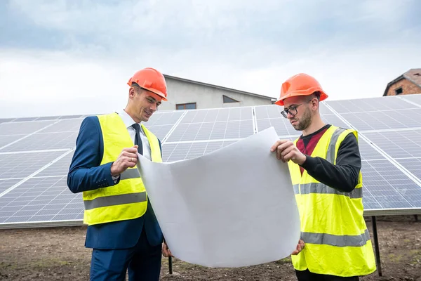 professionals in men's clothing and helmets stand with a tablet standing near the newly installed solar panels. The concept of green electricity in nature
