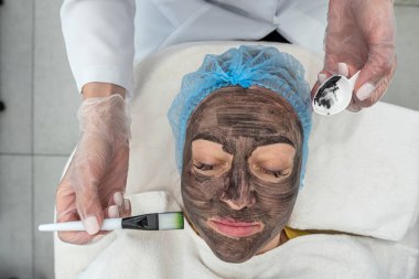 Kozmetik maskeli bir kadının spa merkezindeki yatağında yatışı. Kömürle yüz maskesi soymak. Spa prosedürleri. Cilt bakımı. Güzel bir müşteri, profesyonellerden prosedür alır.