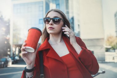 A woman in a formal black suit enjoying a sunny day and walking near the skyscrapers during a work break with a drink in her hand. The concept of the work of a woman in the office
