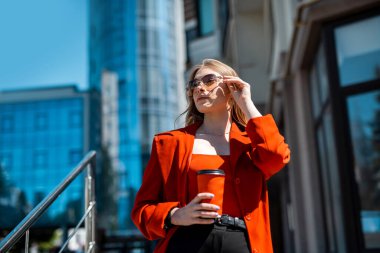 A woman in a formal black suit enjoying a sunny day and walking near the skyscrapers during a work break with a drink in her hand. The concept of the work of a woman in the office