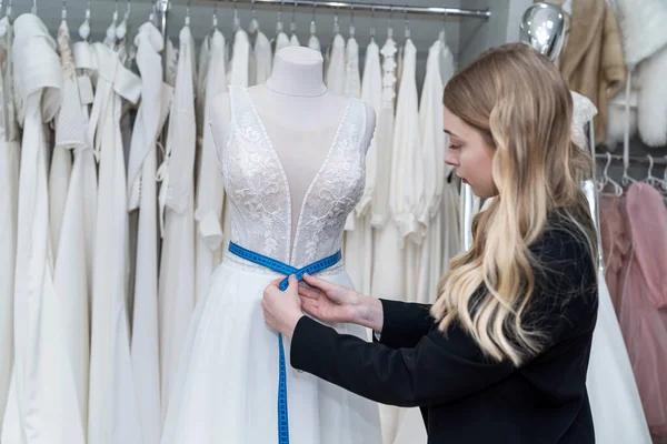 female tailor measuring elegant wedding dress on mannequin using tape measure in store