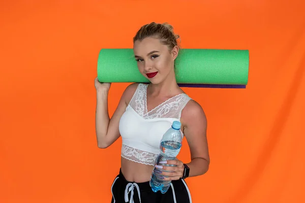 a pretty young well-groomed girl with makeup with a beautiful figure holds a mat and a water bottle near the wall. Healthy lifestyle concept
