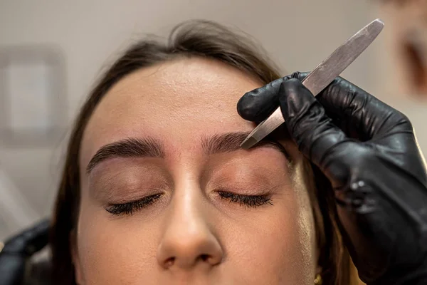 lashmaker makes a beautiful eyebrow contour with tweezers. female face close-up. A beautician does an eyebrow procedure in a beauty salon for a girl. Beauty industry concept.