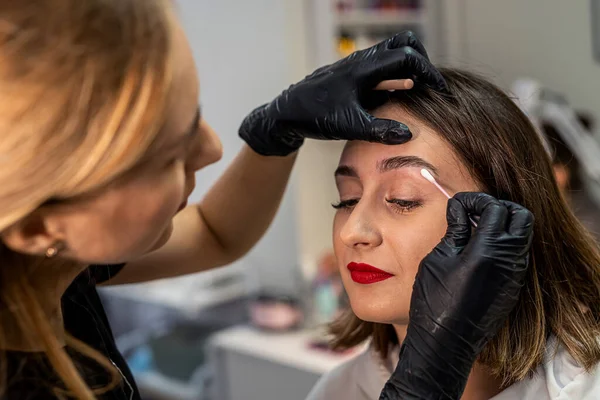 Close-up of an eyebrow designer correcting and touching up eyebrows. A model with dark hair sits in the studio for eyebrow correction by an eyebrow specialist. Makeup and cosmetology concept.