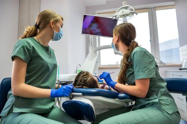 Diş hekimi ve asistanı çocuğun dişlerini bir kamerayla inceler ve resmi ekranda gösterirler. Bir çocuğu dişçide muayene etme konsepti