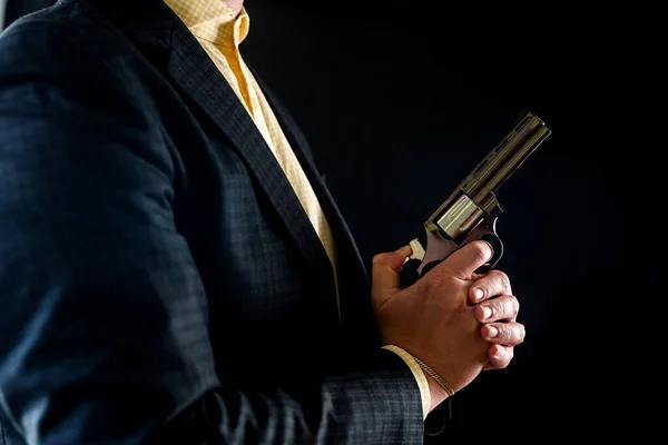 man in a tuxedo and shirt without a face holds a revolver in his hands near his chest isolated on a black background. male hands with a gun. hands in front of the chest with a revolver