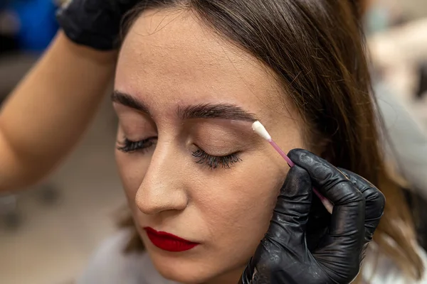 Close-up of an eyebrow designer correcting and touching up eyebrows. A model with dark hair sits in the studio for eyebrow correction by an eyebrow specialist. Makeup and cosmetology concept.