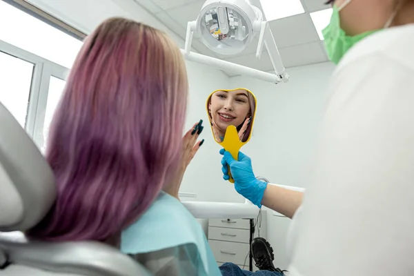 happy female patient is enjoying her beautiful toothy smile looking in the mirror in the dental office held by the dentist. health concept. teeth smile.