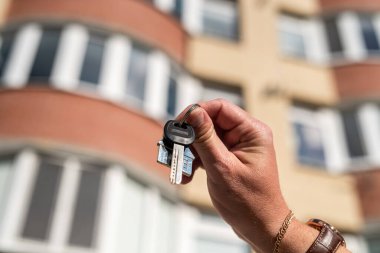 Güçlü erkek, evin anahtarlarını ve anahtarlığı yeni evin önünde yakın çekim şeklinde tutuyor. Gayrimenkul kavramı. Anahtarlı emlakçı