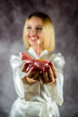 Çekici, heyecanlı, makyajlı ve bornozlu, elinde mumla doğum günü pastası tutan genç bir kadın. Sade bir arka planda izole edilmiş. Bornozla dogum günü