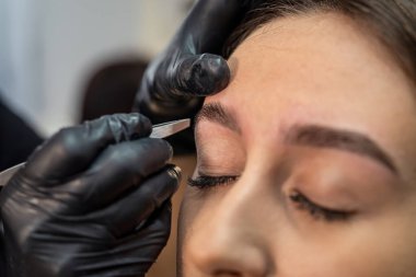 Kirpikçi cımbızla güzel bir kaş çizgisi yapar. Kadın yüzü yakın plan. Güzellik uzmanı bir kız için güzellik salonunda kaş ameliyatı yapıyor. Güzellik endüstrisi konsepti.
