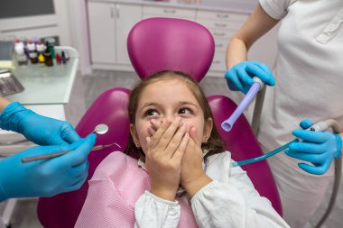 little scared girl covering her mouth with her hands while visiting the dentist. Close-up of an orthodontist trying to check the teeth of a frightened child sitting in a dental chair in a clinic. 