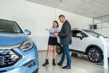 young manager in suit holding clipboard with female client  talking to sales modern car at showroom