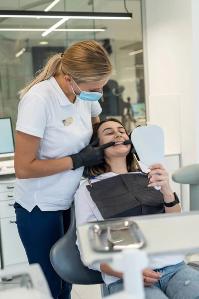 Smiling happy female dentist and girl patient smiling and looking at their white teeth in the mirror while sitting in the dentist\'s office. Concept of teeth whitening and dental implants.