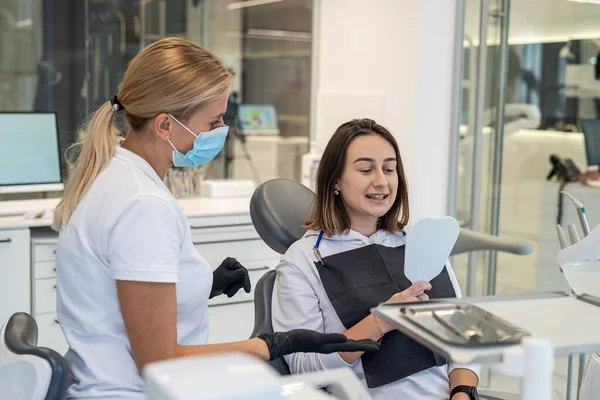 Smiling happy female dentist and girl patient smiling and looking at their white teeth in the mirror while sitting in the dentist\'s office. Concept of teeth whitening and dental implants.