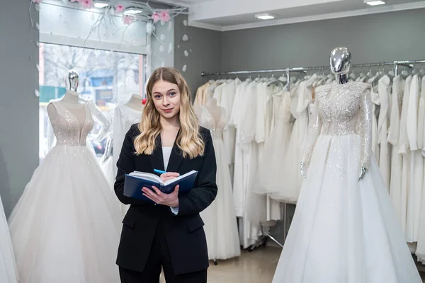Pretty  female dressmaker with diary and pen posing near bridal wear in wedding fashion boutique