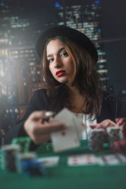 attractive girl in jacket and hat holding poker cards and looking at camera in casino. a woman deals cards. excitement. poker. game for money