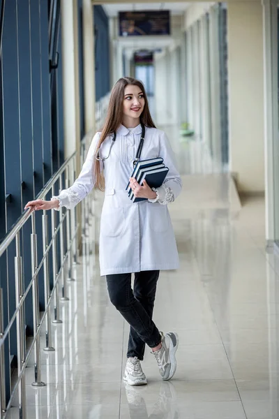 a beautiful student in a medical gown with books in her hands is standing in the corridor of the hospital. Medical student concept. Portrait of a girl