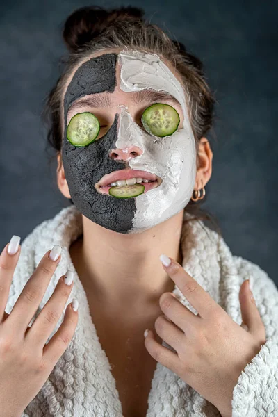 Young woman with facial clay black and white mask holding cucumber slice isolated on dark. Healthy beauty treatment, spa procedure