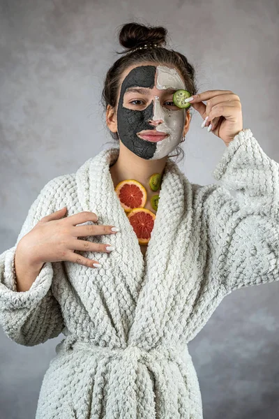 Yüzünde siyah beyaz kilden bir maske olan genç bir kadın taze kivi greyfurt meyvesi tutuyor. Kişisel bakım yaşam tarzı konsepti. Kozmetik insan prosedürü