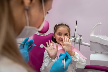 cute little girl is sitting on the dental chair and does not want to treat her teeth by showing appropriate gestures. concept of fear of dentists. healthy oral cavity