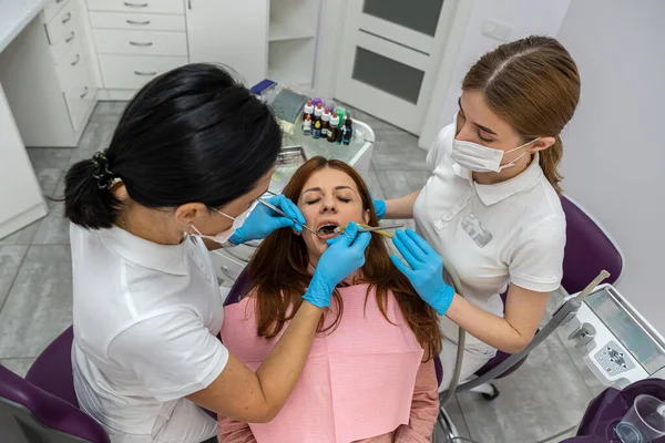female dentist and a female assistant in a special uniform treat the teeth of a female patient. A dentist with an assistant at work in a dental office. Concept of dentistry and healthcare.